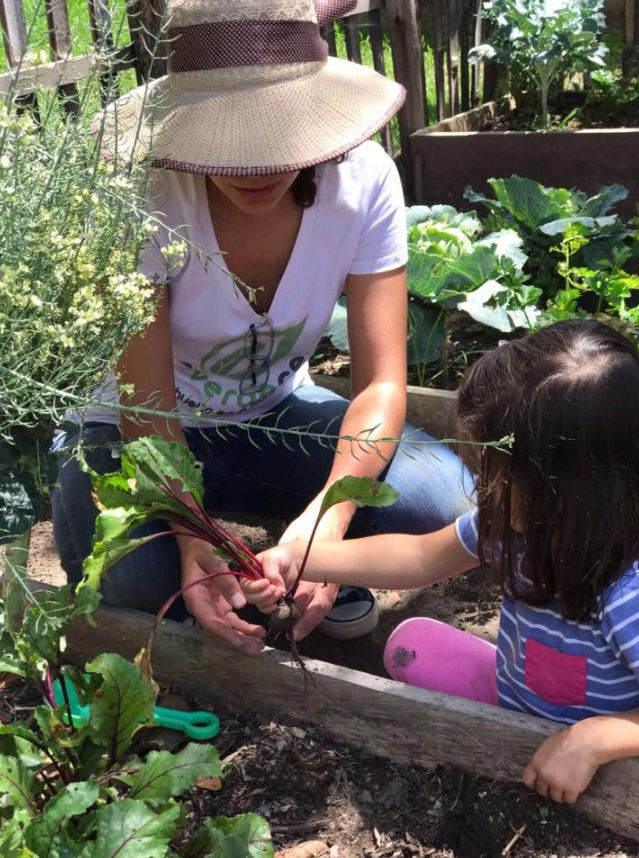 Daniela acompaña a una niña mientras observan y cuidan plantas en un huerto